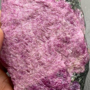 May include: A rough-cut, natural ruby specimen. The stone displays a vibrant pinkish-red hue with areas of darker, purplish-red. The top edge has a dark gray and green coloration. The surface has a textured, crystalline appearance.