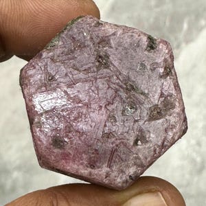 Può includere: Una gemma di rubino grezza, non tagliata, di forma esagonale. Il rubino mostra una tonalità rosso-rosata intensa con inclusioni naturali visibili e una superficie leggermente strutturata. La gemma è tenuta tra le dita, mostrando la sua bellezza naturale.