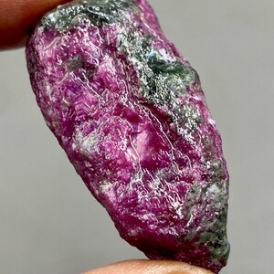 Peut inclure: Un cristal de rubis rose brut avec des inclusions vertes. Le cristal mesure environ 5 cm de long et a une extrémité pointue.