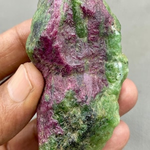 Puede incluir: Una gema en bruto con una mezcla de colores. La piedra tiene un tono magenta intenso, con parches verdes y negros. La superficie es texturizada e irregular, con estructuras cristalinas visibles. La forma general es irregular y orgánica.