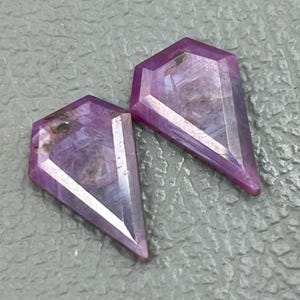May include: Two purple gemstone cabochons cut in a kite shape. The gemstones are faceted and have a translucent appearance.