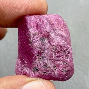 May include: A raw ruby gemstone, displaying a vivid pink colour. The surface of the stone has a textured appearance with dark inclusions. The gemstone is held between two fingers, highlighting its natural, unpolished state. Ideal for jewellery making.