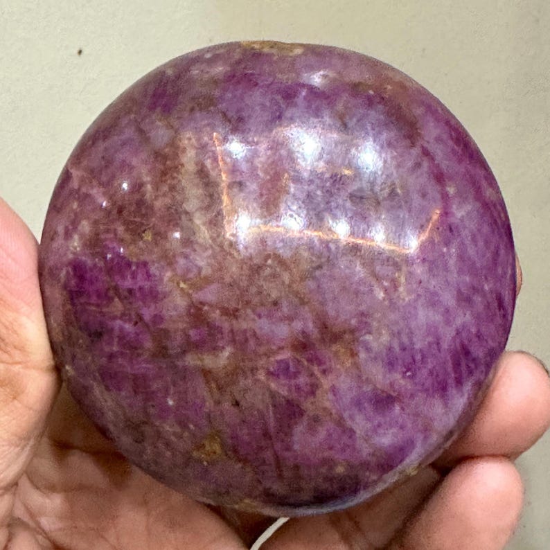 May include: A polished, spherical ruby gemstone displaying a deep magenta and purple hue with natural internal veining and crystalline inclusions throughout the surface. The stone has a smooth, rounded texture and a reflective, glossy finish.