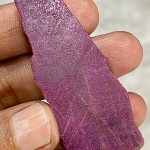 Puede incluir: Una gema en bruto, alargada, de color magenta intenso. La piedra tiene una superficie texturizada con estrías visibles y un borde ligeramente más claro. La gema se sostiene en una mano, mostrando su forma natural y en bruto.