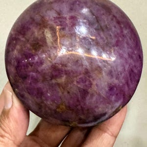 May include: A polished, spherical gemstone featuring a deep magenta and purple colour palette with natural internal veining and mottled patterns. The surface is smooth and reflective, showcasing variations in opacity and mineral inclusions throughout.