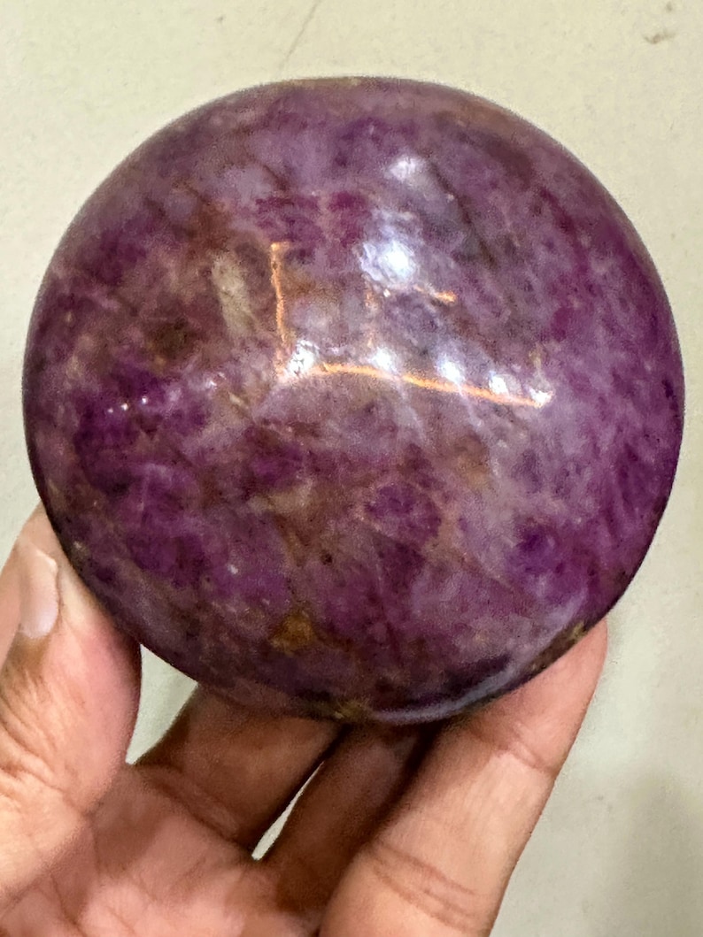 May include: A polished, spherical gemstone featuring a deep magenta and purple colour palette with natural internal veining and mottled patterns. The surface is smooth and reflective, showcasing variations in opacity and mineral inclusions throughout.