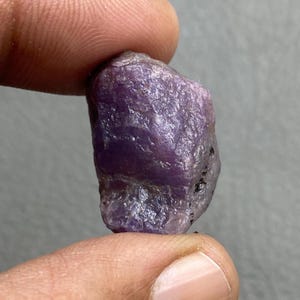 May include: A rough, uncut purple gemstone with a textured surface. The stone is held between two fingers, showcasing its natural, irregular shape and color variations. The gemstone has a deep, rich purple hue.