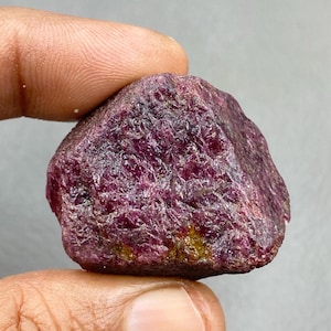Può includere: Una gemma di rubino grezza, non tagliata, di colore rosso-viola intenso. La superficie è ruvida e irregolare, con piccole aree gialle visibili. La gemma è tenuta tra due dita su uno sfondo grigio neutro. Esemplare minerale grezzo.