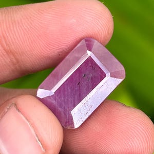 May include: A pink gemstone with a rectangular cut and a faceted finish. The gemstone is held in a hand.