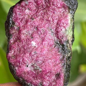 Puede incluir: Primer plano de una gema en bruto sin cortar. La piedra tiene un centro rosa vibrante con una superficie texturizada, rodeada por una capa exterior oscura, casi negra. La piedra natural se sostiene entre los dedos.