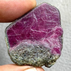Puede incluir: Una gema de rubí en bruto, de un tono magenta intenso. La piedra tiene una superficie texturizada con áreas grises y verdes. El rubí se sostiene entre los dedos, mostrando su estado natural y sin pulir.