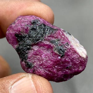 May include: A rough, pink ruby gemstone with black inclusions. The ruby is a natural, unpolished stone.
