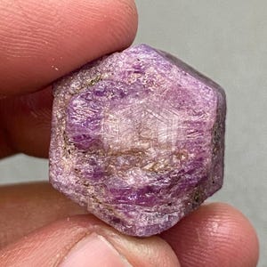 May include: A raw ruby gemstone, hexagonal in shape, with a deep pinkish-purple colour. The gem features lighter, crystalline areas and a surface with natural textures and colour variations. Some areas appear more opaque.