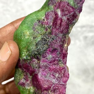Puede incluir: Un rubí en bruto en zoisita. La piedra presenta una vibrante mezcla de cristales de rubí rojo intenso y zoisita verde, con algunas inclusiones negras. La gema natural se sostiene en una mano.