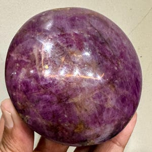May include: A polished, spherical gemstone with a deep magenta and purple hue, featuring natural internal crystalline structures, cloudy white inclusions, and dark mineral veins throughout its smooth, reflective, and rounded surface.