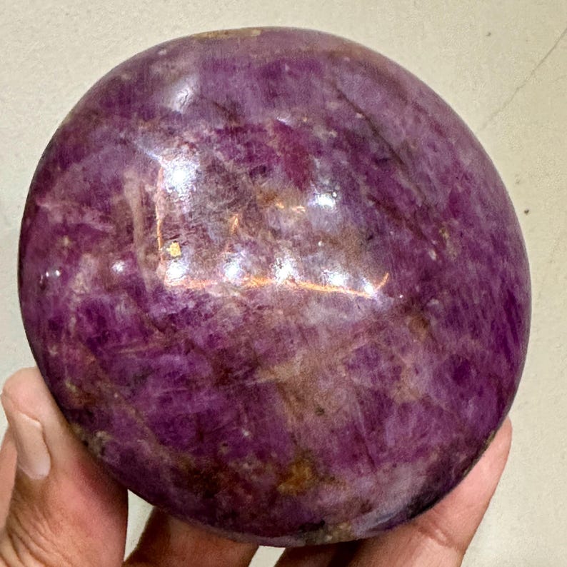 May include: A polished, spherical gemstone with a deep magenta and purple hue, featuring natural internal crystalline structures, cloudy white inclusions, and dark mineral veins throughout its smooth, reflective, and rounded surface.