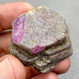 May include: A rough, uncut ruby gemstone with a mix of gray and brown tones. The ruby has vibrant pink and red crystalline inclusions. The stone's surface is textured, showcasing natural formations. This raw gemstone is ideal for collectors or jewelry makers.