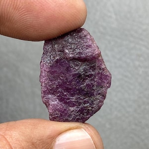 May include: A raw, uncut ruby gemstone held between fingers. The ruby has a deep, rich purple colour with a textured surface. The natural gemstone is irregularly shaped, showcasing its raw, unpolished state.