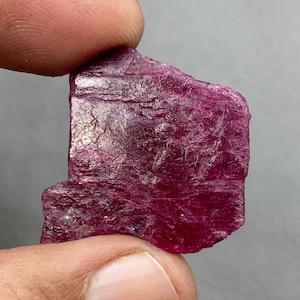 May include: A raw, uncut ruby gemstone in a deep magenta hue. The natural mineral has a textured surface. The stone is held between two fingers, highlighting its natural form and potential for jewellery or collection.