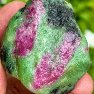 May include: A natural gemstone with a mix of colours. The stone has a light green base, with patches of deep pink and black. The rough, unpolished texture of the stone is visible. The gemstone is a natural, unrefined product.