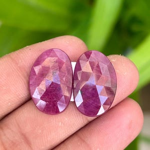 Puede incluir: Dos piedras preciosas de rubí de forma ovalada y facetadas, de un tono rosa intenso. Las piedras están colocadas sobre un fondo blanco.