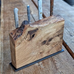 Könnte beinhalten: Ein rechteckiger Messerblock aus einer dicken Scheibe naturfarbenem, hellbraunem Holz mit sichtbarer Maserung, Astlöchern und einer Naturkante. Der Block steht auf einem dünnen, flachen schwarzen Metallsockel und enthält vier Küchenmesser.