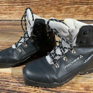 Op de afbeelding: Paar zwarte en grijze Fischer skischoenen met zwart-witte veters. De laarzen hebben een high-top ontwerp met het Fischer logo aan de zijkant. De laarzen staan op een houten ondergrond.