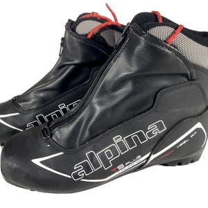 Peut inclure: Paire de chaussures de ski de fond Alpina TS Plus noires. Les chaussures ont une fermeture éclair sur le devant, des accents blancs et des lacets rouges. Les mots "alpina" et "HEEL FIT" sont visibles.