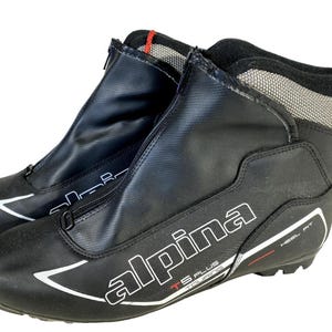 Peut inclure: Paire de chaussures de ski Alpina TS Plus Touring noires. Les chaussures ont une fermeture éclair sur le devant, des accents blancs et le nom de la marque est bien en évidence. Les chaussures sont conçues pour un ajustement au talon et ont un accent rouge sur le côté.