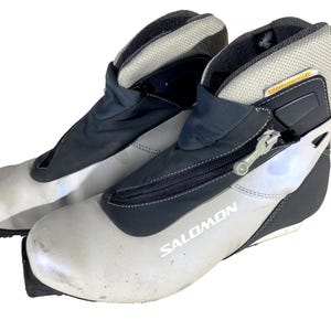 Puede incluir: Par de botas de esquí de fondo Salomon. Las botas son principalmente blancas con detalles en negro y gris. Cuentan con cierre de cremallera y el logotipo de Salomon. Diseñadas para deportes de invierno.