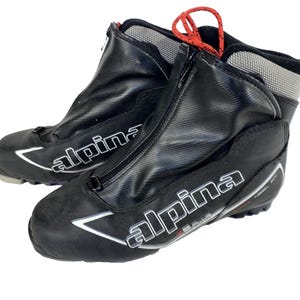 Peut inclure: Paire de chaussures de ski de fond Alpina noires. Les chaussures ont une fermeture éclair sur le devant, des accents blancs et des lacets rouges. Le nom de la marque "Alpina" est affiché en blanc sur le côté de chaque chaussure.