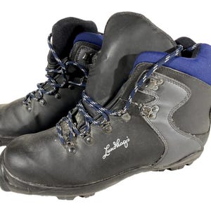 Puede incluir: Par de botas de senderismo negras y grises con cordones azules y blancos. Las botas tienen un diseño de caña alta con ojales metálicos y la marca "Lundhags" en escritura blanca.