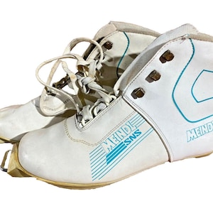 Puede incluir: Par de botas de esquí blancas vintage con detalles azules y la marca "MEINDL". Las botas tienen cordones y herrajes metálicos. Diseñadas para esquí de fondo.