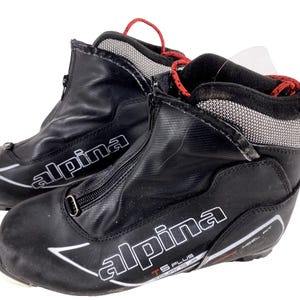 Peut inclure: Paire de chaussures de ski de fond noires avec la marque "Alpina" en blanc. Les chaussures ont une fermeture éclair sur le devant, des lacets rouges et un col en mesh. Elles sont conçues pour le ski de fond et portent l'inscription "TS PLUS".