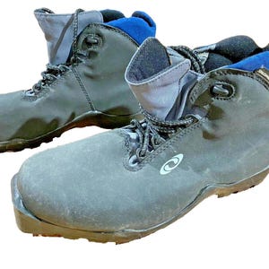 Puede incluir: Un par de botas de senderismo grises y negras con detalles azules. Las botas tienen un diseño con cordones y un logotipo en el lateral. Las suelas son marrones. Estas botas están diseñadas para actividades al aire libre.