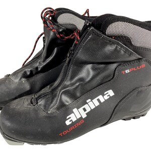 Peut inclure: Paire de chaussures de ski de randonnée Alpina T5 Plus noires. Les chaussures ont un extérieur noir avec une fermeture éclair, des lacets rouges et noirs, et le logo Alpina en blanc. Les chaussures sont conçues pour le ski de fond.
