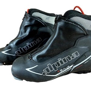 Peut inclure: Paire de chaussures de ski de fond Alpina TS Plus Touring JR noires. Les chaussures ont une fermeture éclair sur le devant, des accents blancs et le nom de la marque en blanc. La zone du talon a une tirette rouge et les mots "HEEL FIT".