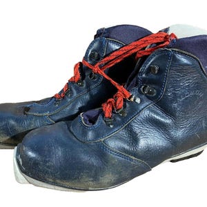 Puede incluir: Par de botas de esquí azul marino vintage con cordones rojos. Las botas tienen un diseño de caña alta con ojales metálicos y suela blanca. Las botas muestran signos de desgaste. Un clásico del equipamiento de deportes de invierno.