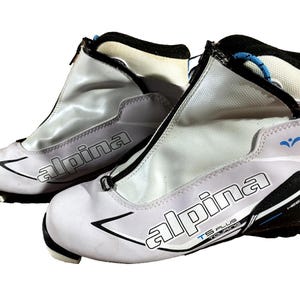 Peut inclure: Paire de chaussures de ski de fond Alpina TS Plus blanches et grises. Les chaussures ont des fermetures éclair noires, des accents noirs et bleus, et le logo Alpina en noir. Les chaussures sont conçues pour le touring et ont un design de talon.