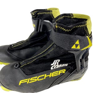 Op de afbeelding: Paar zwarte en gele Fischer JR Combi langlauf skischoenen. De schoenen hebben een zwart bovenwerk met gele stiksels en accenten, inclusief het Fischer-logo. De schoenen hebben een ritssluiting en een klittenband.