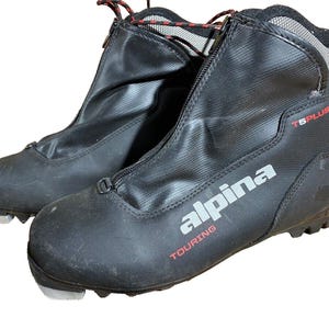 Peut inclure: Paire de chaussures de ski Alpina Touring noires. Les chaussures ont une fermeture éclair sur le devant, le nom de la marque "alpina" en argent et le mot "TOURING" en rouge. Les bottes sont étiquetées "T5 PLUS" et ont un détail de lacets noirs et rouges.