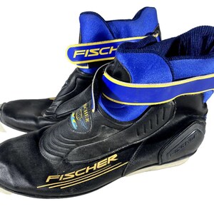 Puede incluir: Par de botas de esquí de fondo FISCHER negras y azules. Las botas tienen una base negra con detalles azules en el cuello y las correas. El logotipo de FISCHER es visible en letras doradas. Las botas están diseñadas para deportes de invierno.