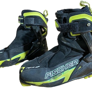 Puede incluir: Par de botas de esquí Fischer RCS negras y amarillo neón. Las botas tienen un diseño de caña alta con correas ajustables y cierre de cremallera. El logotipo de Fischer es visible en el lateral de la bota.