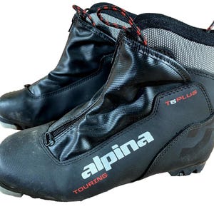 Peut inclure: Paire de chaussures de ski de randonnée Alpina T5 Plus noires. Les chaussures ont une fermeture éclair sur le devant, des lacets rouges et noirs et le logo Alpina en blanc. Conçues pour le ski de fond.