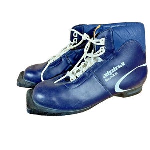 May include: A pair of navy blue Alpina Blaze ski boots. The boots have white laces and a white curved design on the side. The boots have a black sole and are made of a shiny material.