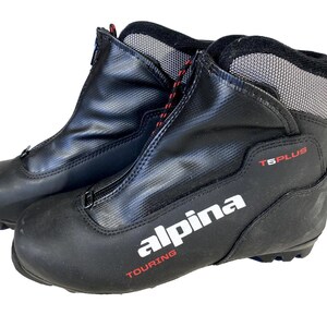 Peut inclure: Paire de chaussures de ski de fond Alpina Touring noires. Les chaussures ont une fermeture éclair sur le devant, des lacets rouges et noirs, et le texte "T5 PLUS" et "TOURING" en rouge. Le logo Alpina est en blanc.