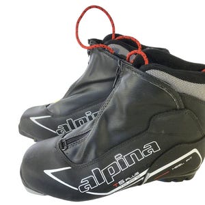 Peut inclure: Paire de chaussures de ski de randonnée Alpina TS Plus noires. Les chaussures ont une fermeture éclair sur le devant, des accents blancs et rouges et des boucles de tirage rouges. La marque "Alpina" est affichée en blanc sur le côté. Conçues pour le ski de fond.