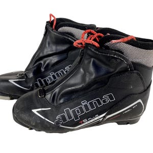 Peut inclure: Paire de chaussures de ski de fond Alpina noires. Les chaussures ont une fermeture éclair sur le devant, des lacets rouges et la marque "Alpina" en blanc. Les chaussures sont conçues pour les sports d'hiver et les activités de plein air.