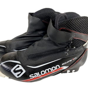 May include: Pair of black and white Salomon Equipe CL cross-country ski boots. The boots feature a zip front, mesh detailing, and red accents. The Salomon logo is displayed on the side.