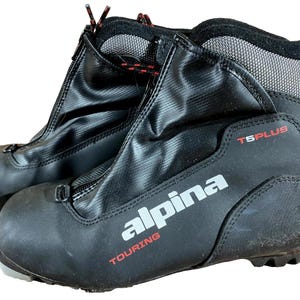 Peut inclure: Paire de chaussures de ski de randonnée Alpina T5 Plus noires. Les chaussures ont une fermeture éclair sur le devant, des lacets rouges et noirs et le logo Alpina en blanc. Conçues pour le ski de fond, elles ont un design robuste et durable.